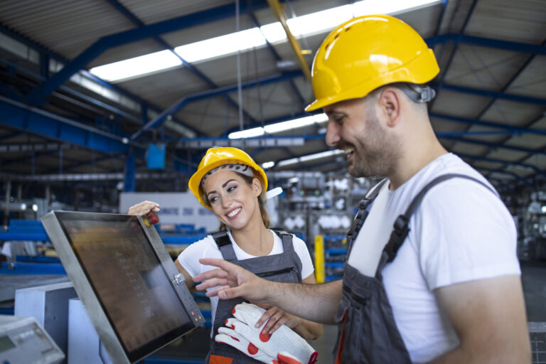 Factory worker explaining trainee how to operate industrial machine using new software on touch screen computer.