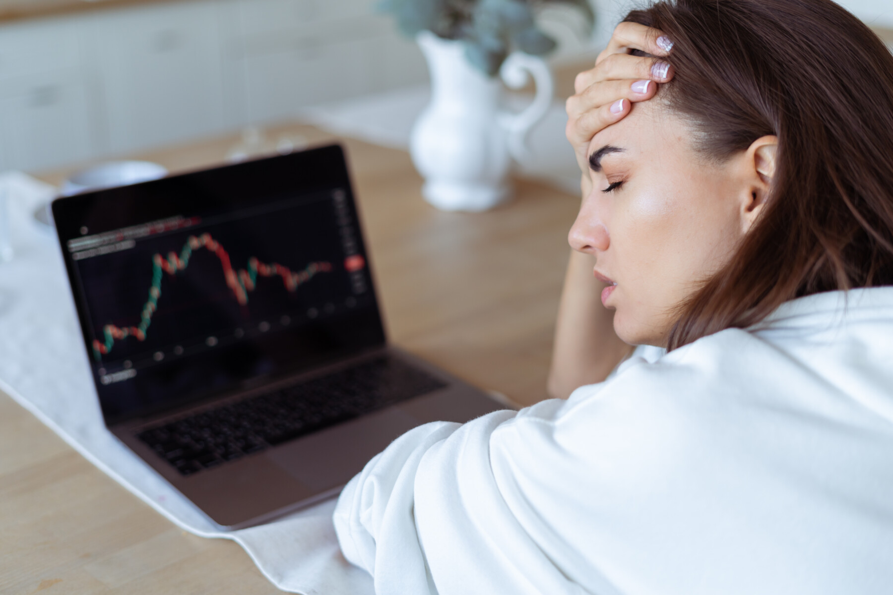 Young woman at home in the kitchen in a white hoodie with a laptop, graph on the screen, upset, unhappy, cryptocurrency falls down