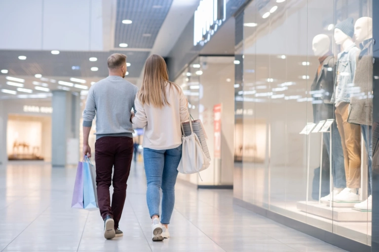 Rear View Young Casual Couple Moving Along Shop Window Mall While Leaving It After Buying What They Wanted
